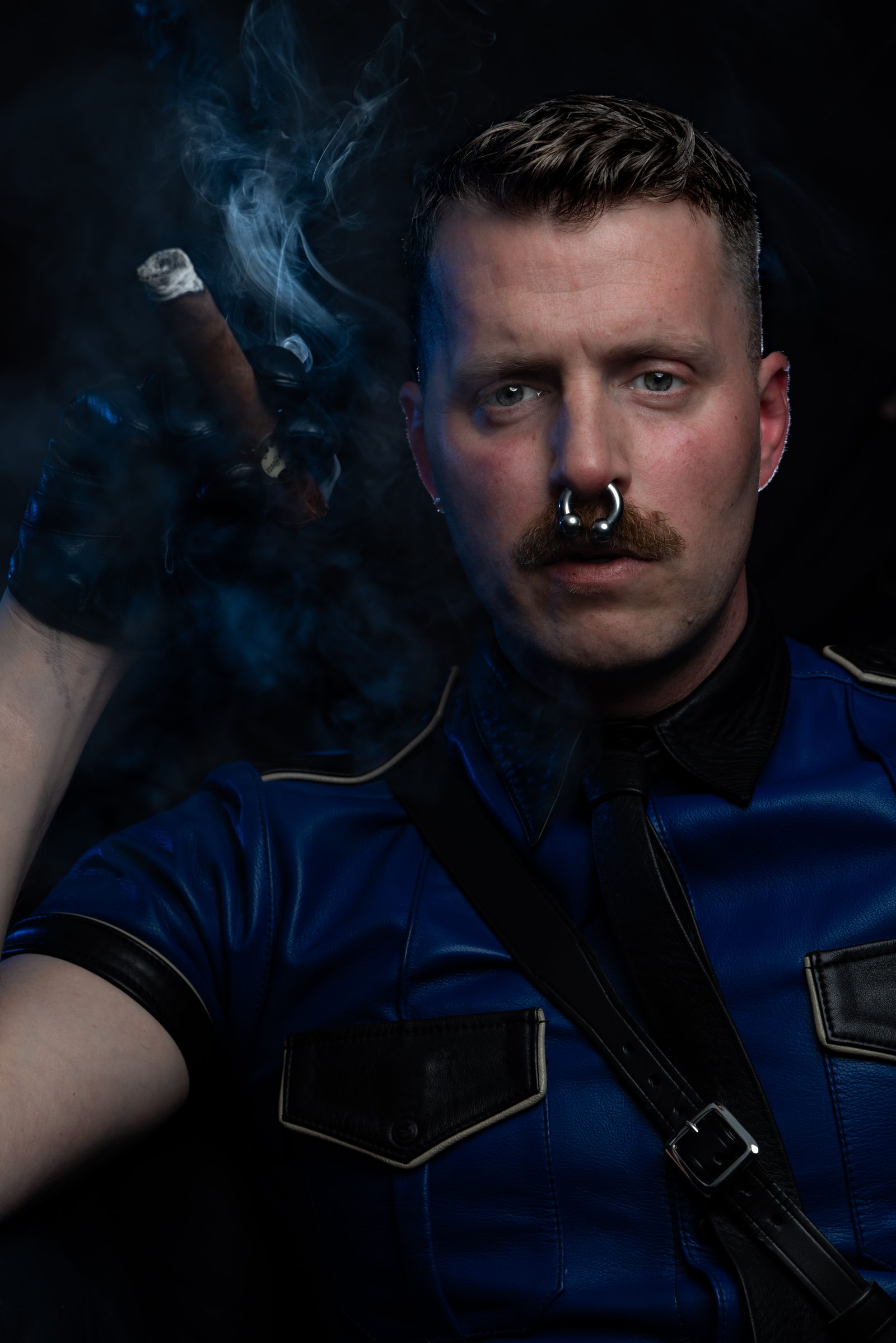 A leather portrait by Nick Leoni, a mustachioed man in a blue leather police shirt raising a cigar through blue smoke, commanding and at ease