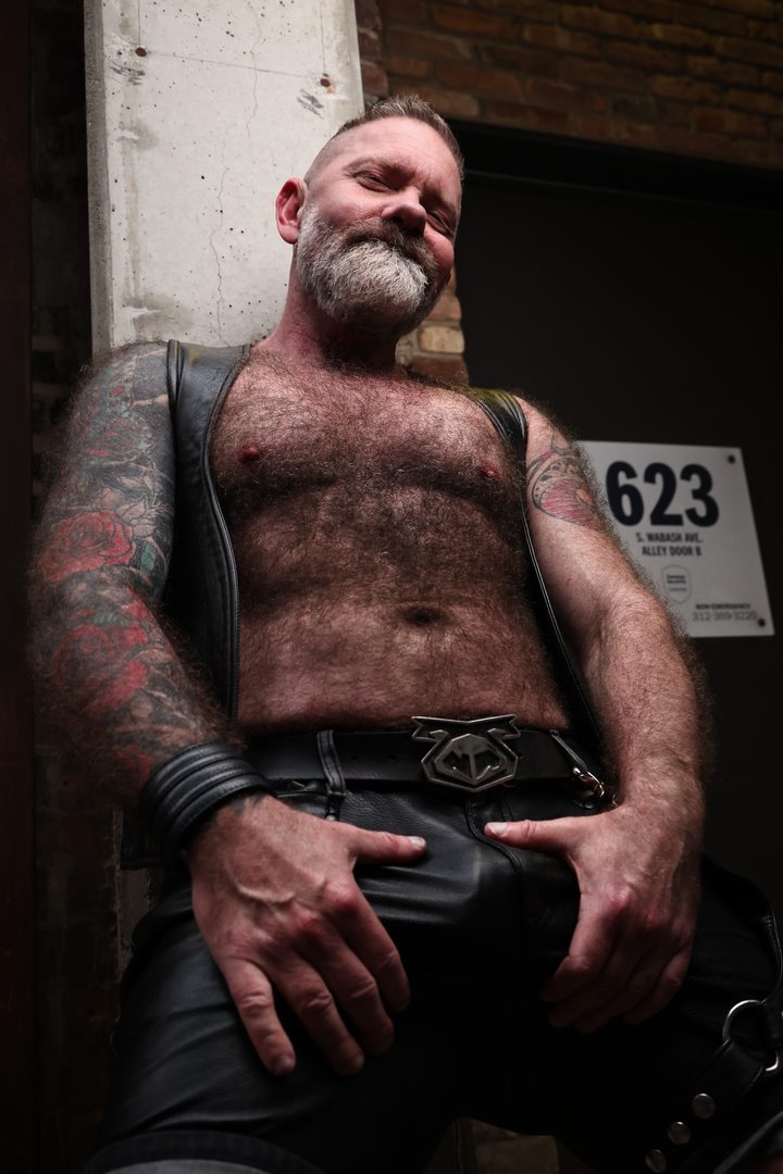 A silver-bearded man in a leather vest and chaps leans into an alley doorway marked 623, tattooed arms framing a calm, self-possessed look.