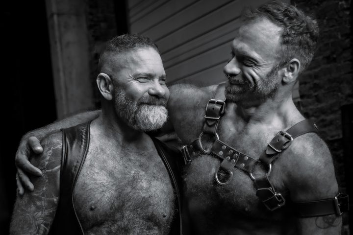 A black-and-white frame of two leather men sharing a laugh, one resting his head toward the other's shoulder in an easy, warm moment.