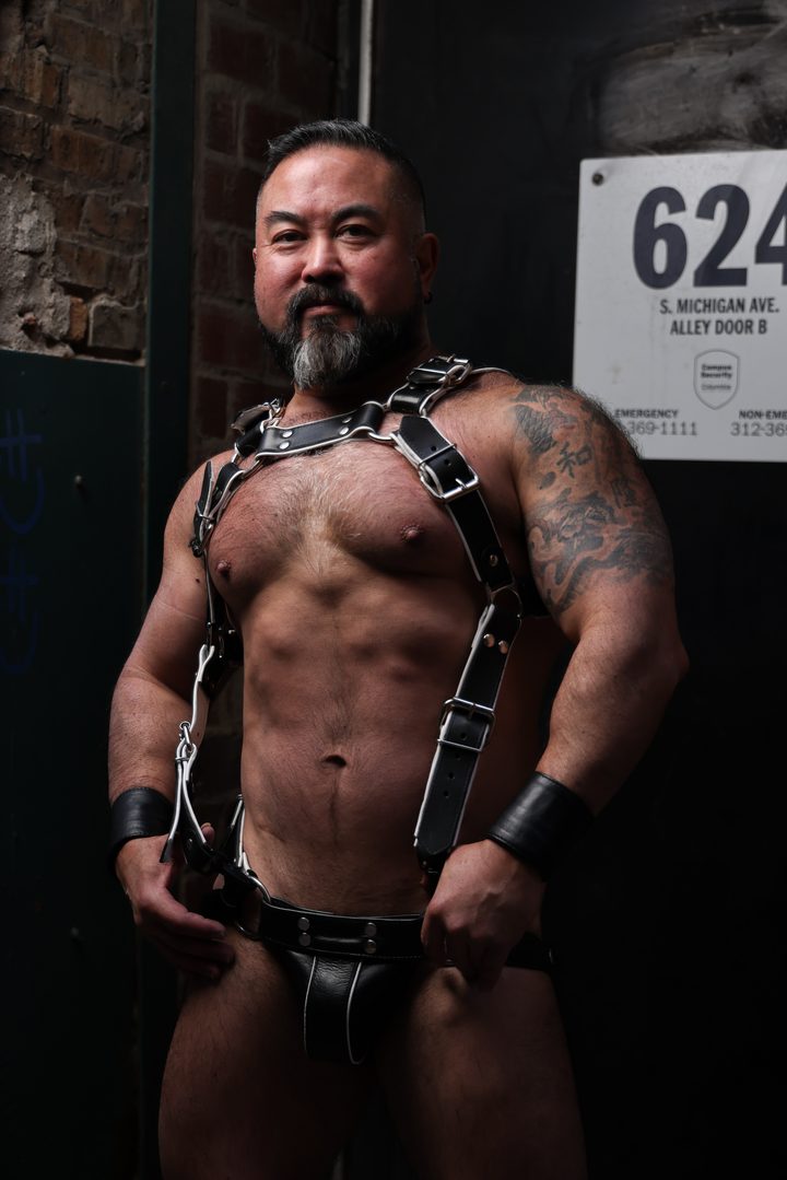 A bearded Asian man in a leather harness and jock stands grounded in a dim alley doorway marked 624, tattooed arm catching a low warm light.