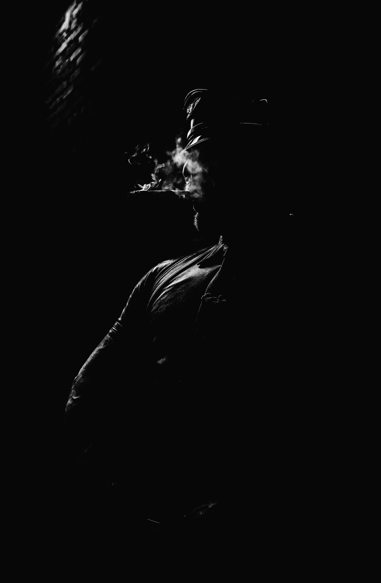 A near-silhouette black-and-white portrait of a capped figure exhaling cigar smoke, the form emerging from deep shadow into a thin edge of light.