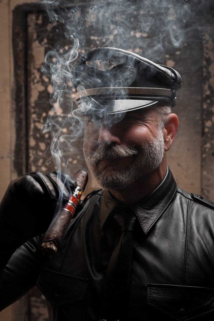 A silver-mustachioed man in a leather uniform shirt, tie, and Muster cap draws on a cigar, the smoke catching warm light against a distressed wall.