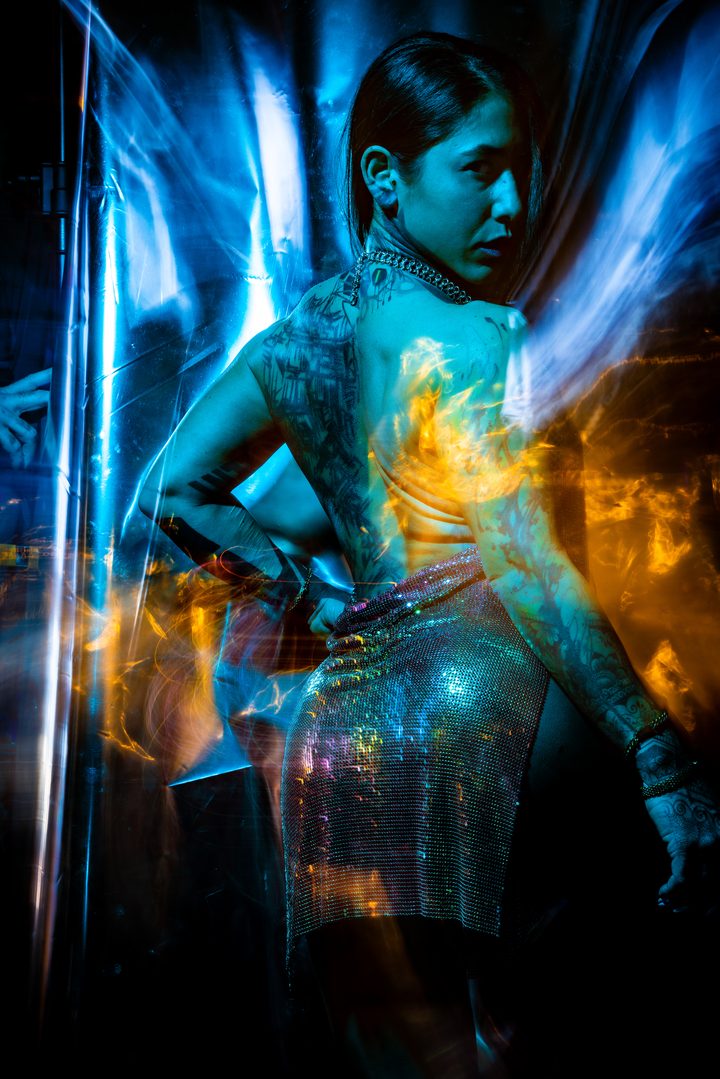 Fire projected onto skin and a chainmail garment, seen through scratched glass in blue and amber, sharp and spectacular.